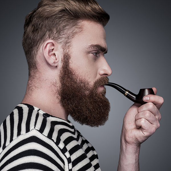 Side view of confident young bearded man in striped clothing smoking a pipe and looking away while standing against grey background